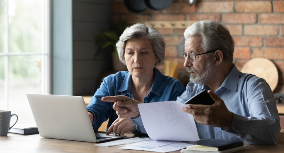 An older couple sitting at their kitchen table looking at a laptop and paperwork
